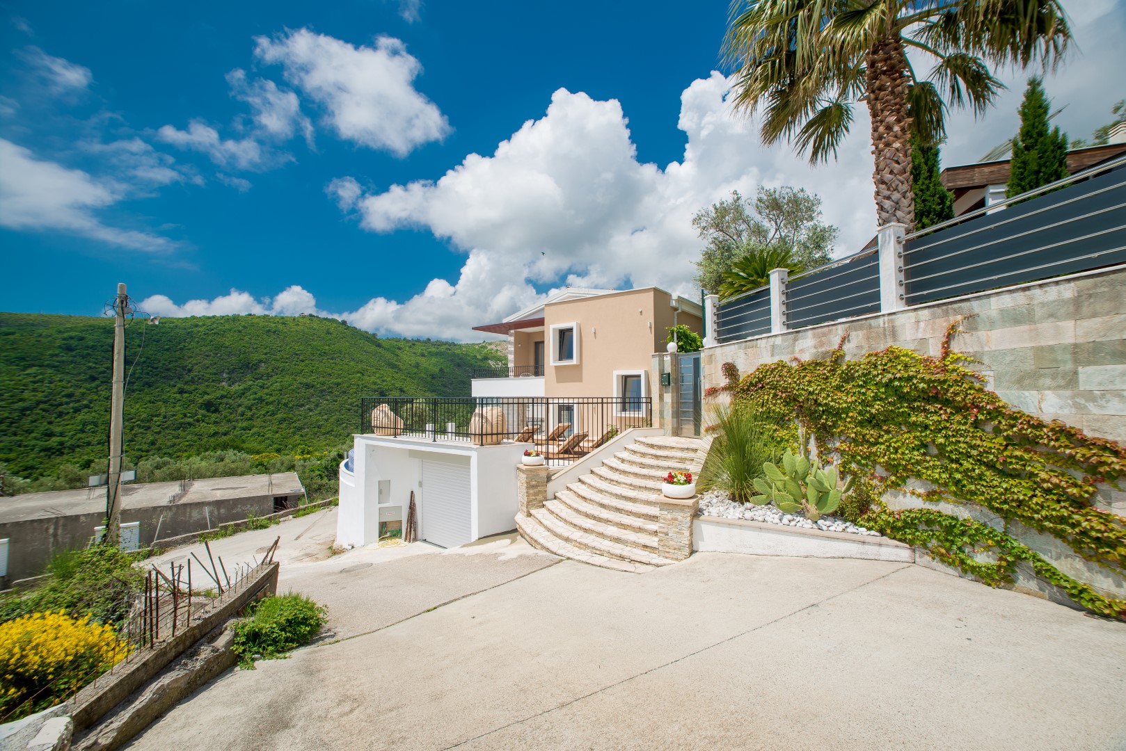 Modern villa in Bečići, Montenegro: facade with a terrace, stone staircase, palm tree, and views of green mountains.