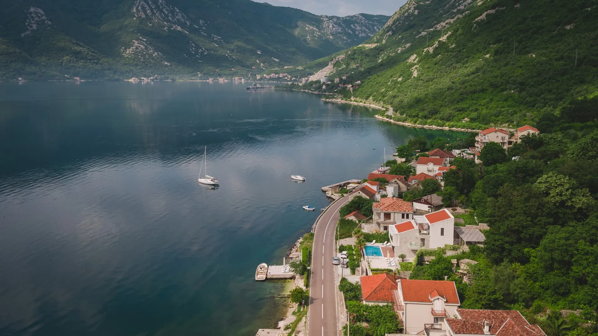Aerial view of a coastal village with houses, a road, and boats on the water surrounded by green mountains.