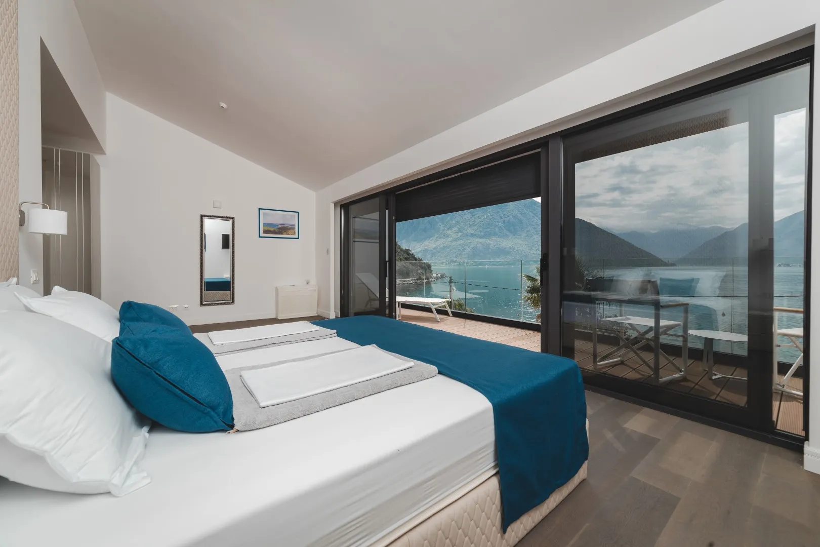 Modern bedroom with a large bed and blue bedding, featuring sliding glass doors leading to a balcony with a lake view.