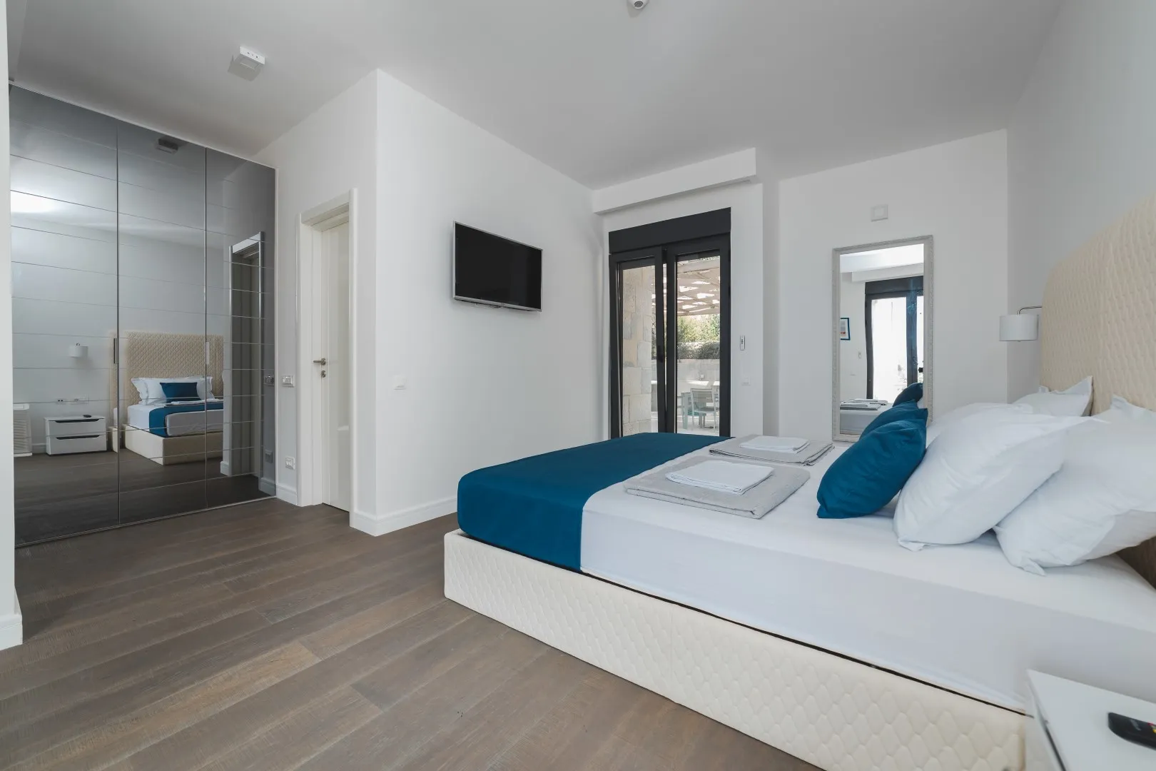 Modern bedroom featuring a large bed with blue bedding, mirrored wardrobe, wall-mounted TV, and wood flooring.