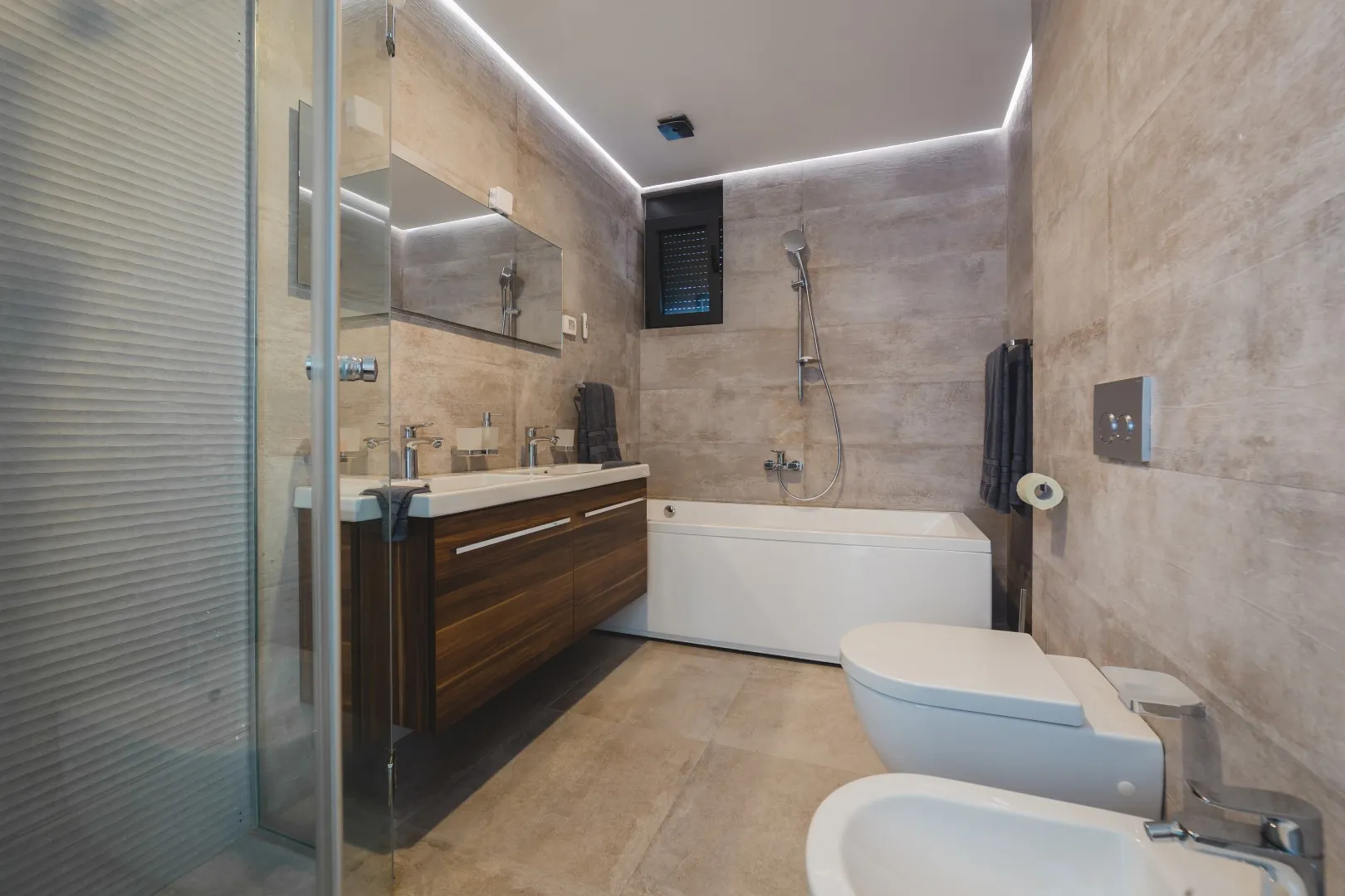 Modern bathroom featuring a wood vanity, white bathtub, wall-mounted toilet, shower system, and beige tiled walls with LED lighting.