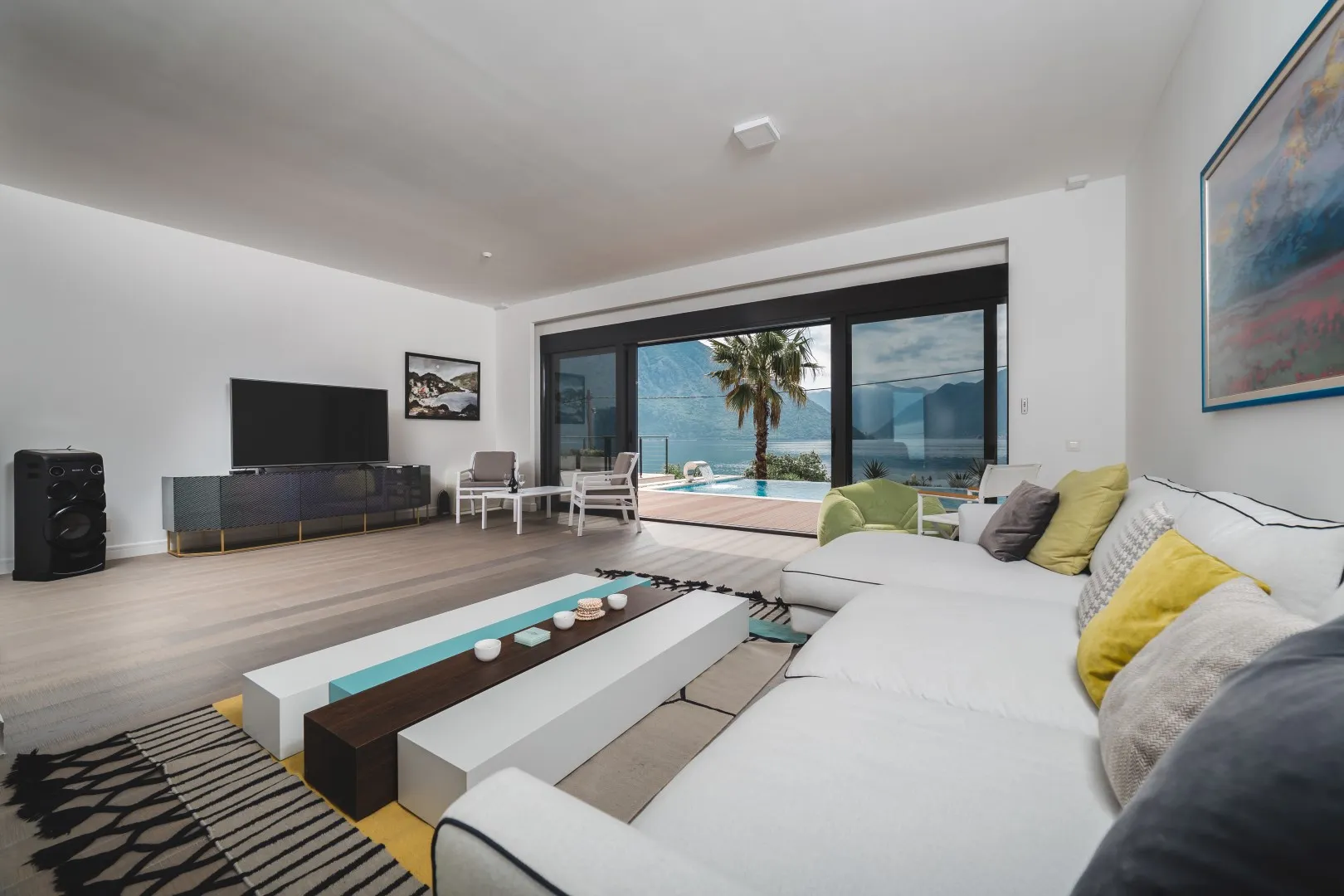 Bright living room with white sofa, TV, and sliding doors opening to a pool and sea view.