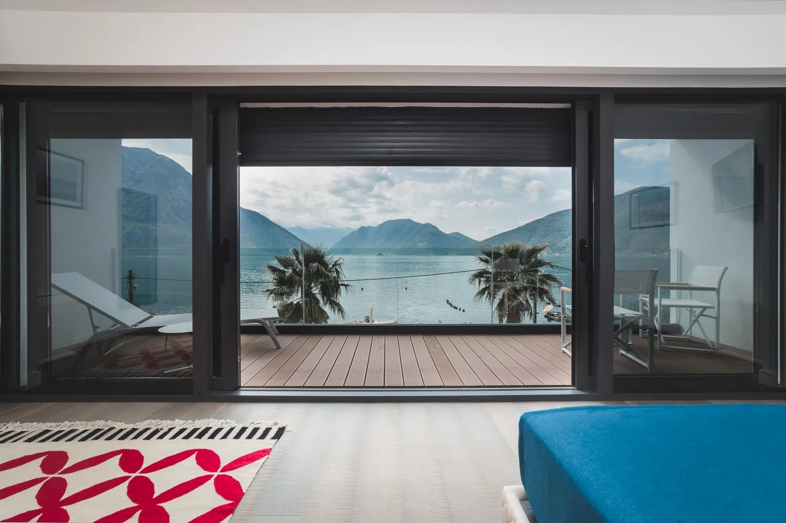 View from an interior room looking out through sliding glass doors onto a wooden balcony overlooking a lake and mountains.