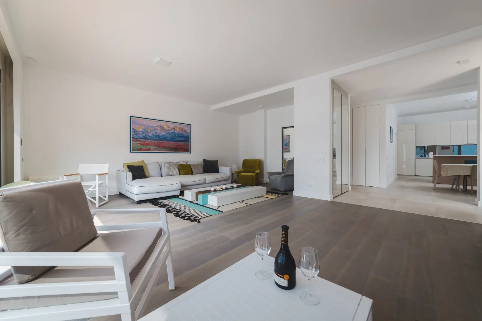 Bright living room with a white sofa, coffee table, wall art, and an open view into the kitchen area.