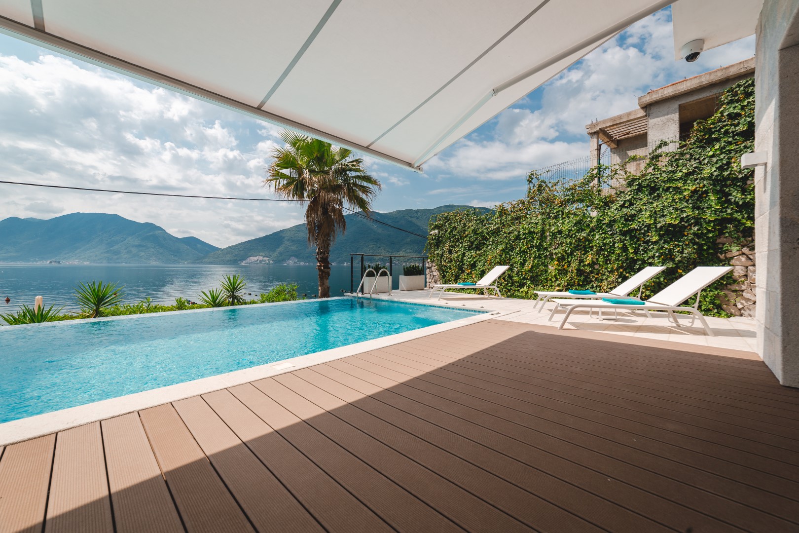 Terrace with a pool and a view of Kotor Bay, villa in Strp, Montenegro: sun loungers, wooden deck, and a palm tree.