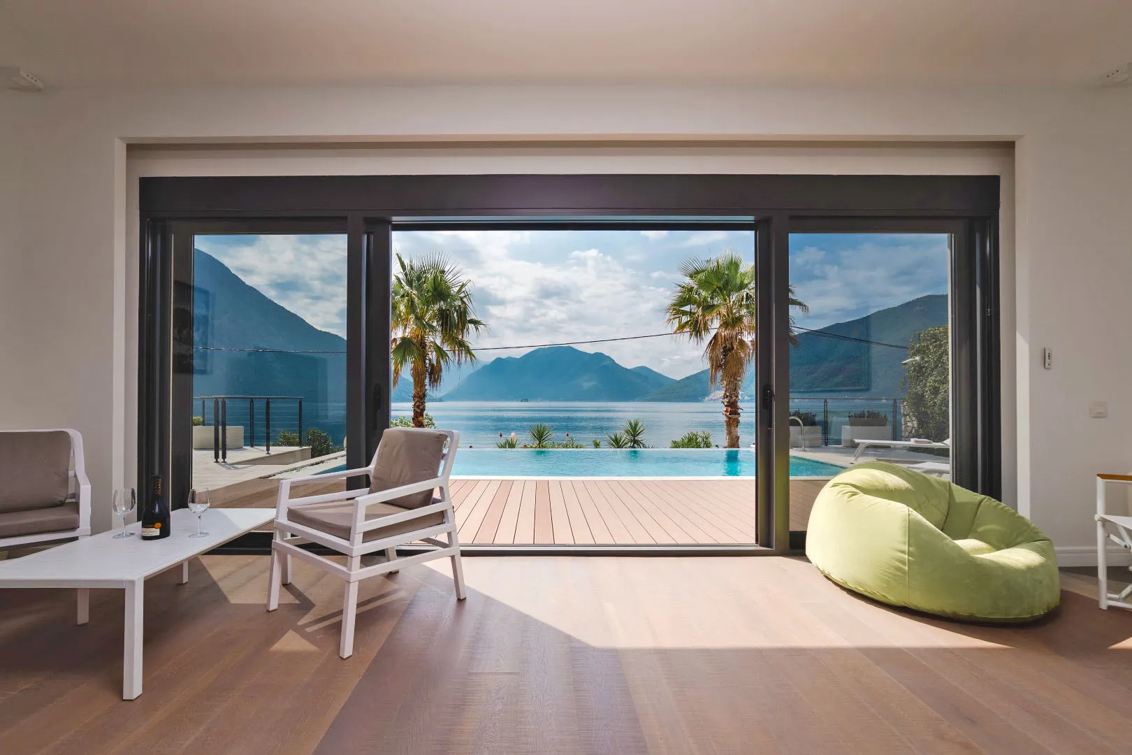 Bright room with wood floors, white chairs, and a green beanbag opening to a deck with a pool and sea view.