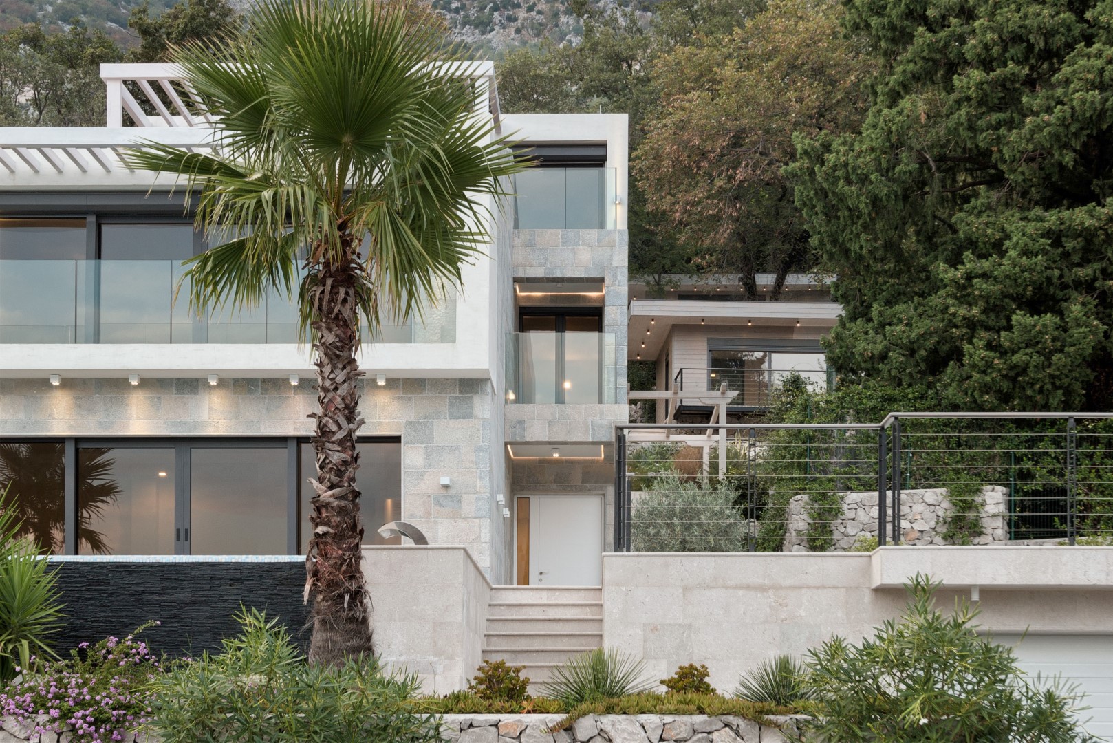 Modern villa in Strp, Montenegro: stone facade, panoramic windows, terrace, and a palm tree against a mountain landscape.