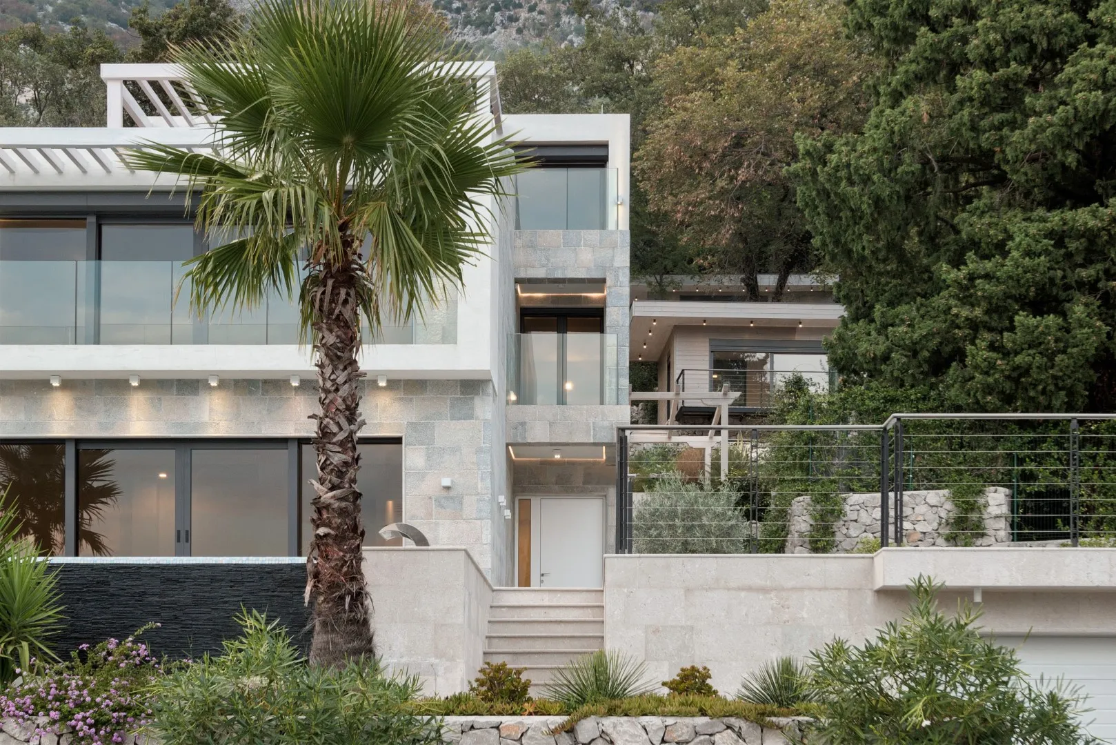 Modern stone house exterior featuring a palm tree, glass balconies, and hillside landscaping.