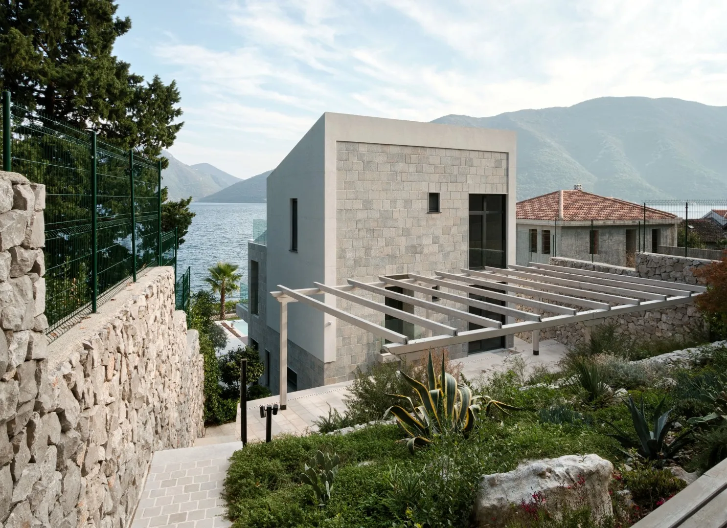 Modern stone house with a pergola and retaining wall overlooking the water and mountains.