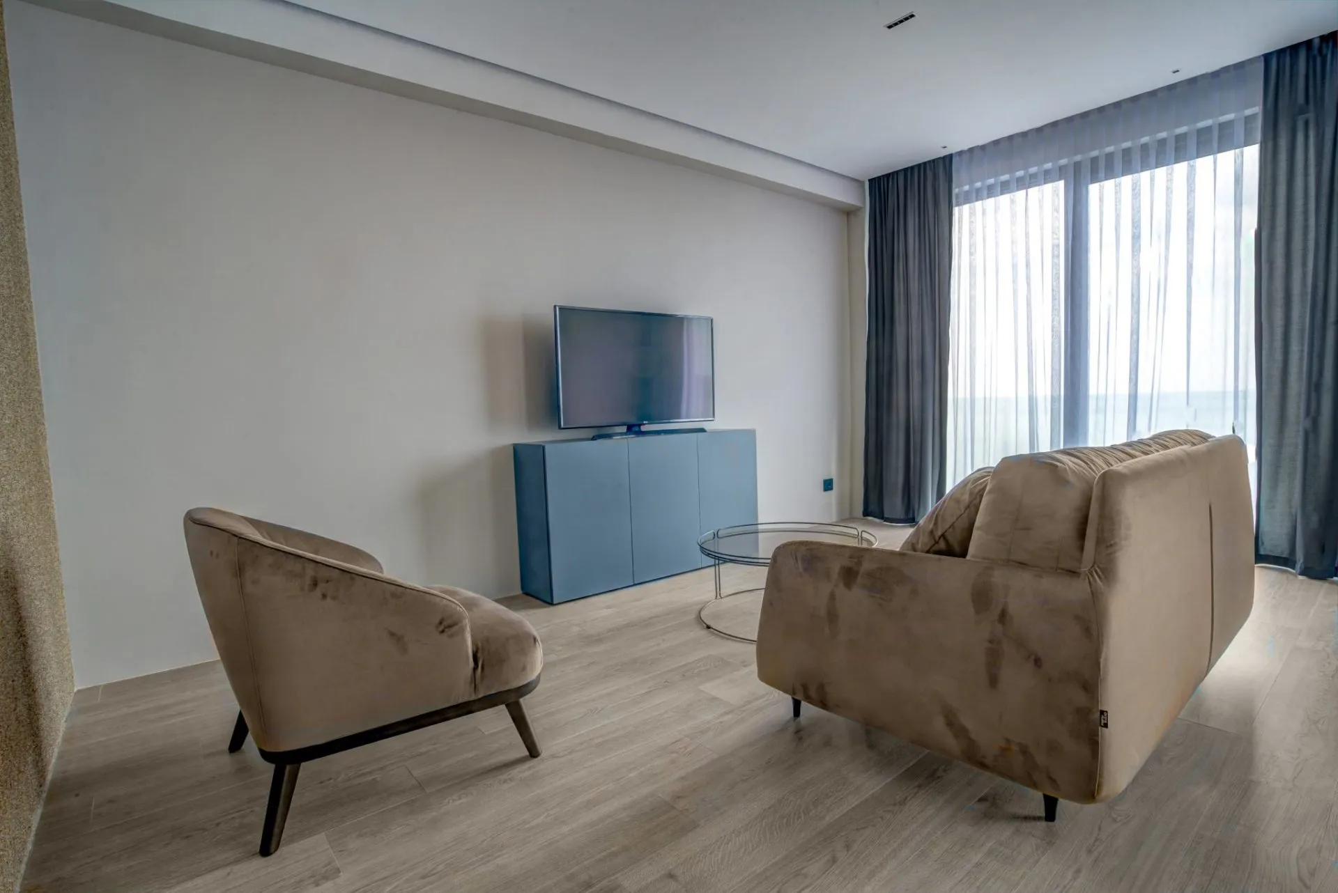 Bright room with beige sofa, armchair, blue cabinet, and TV on light wood flooring near curtains.