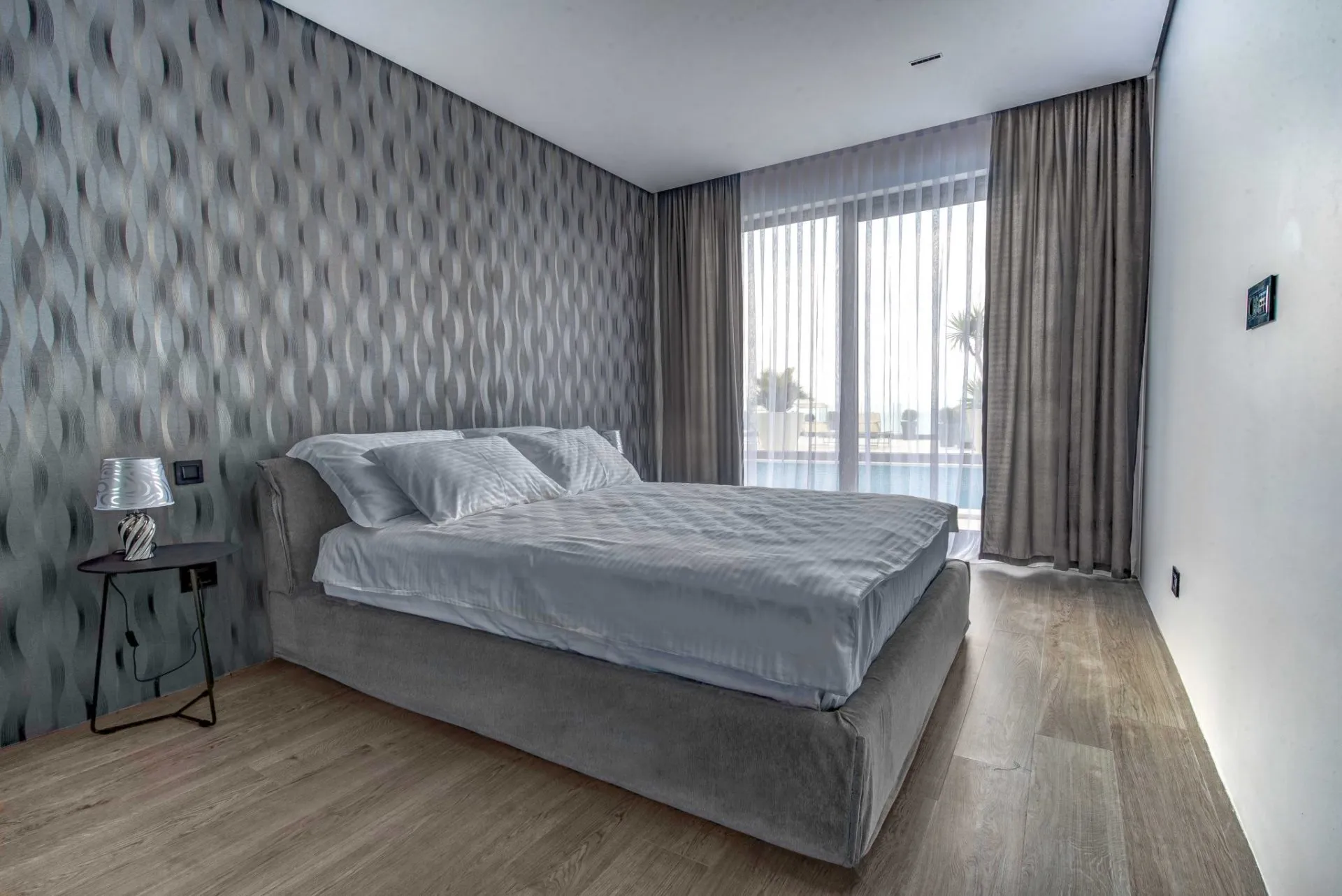 Modern bedroom featuring a grey upholstered bed, patterned wallpaper, and wood flooring.