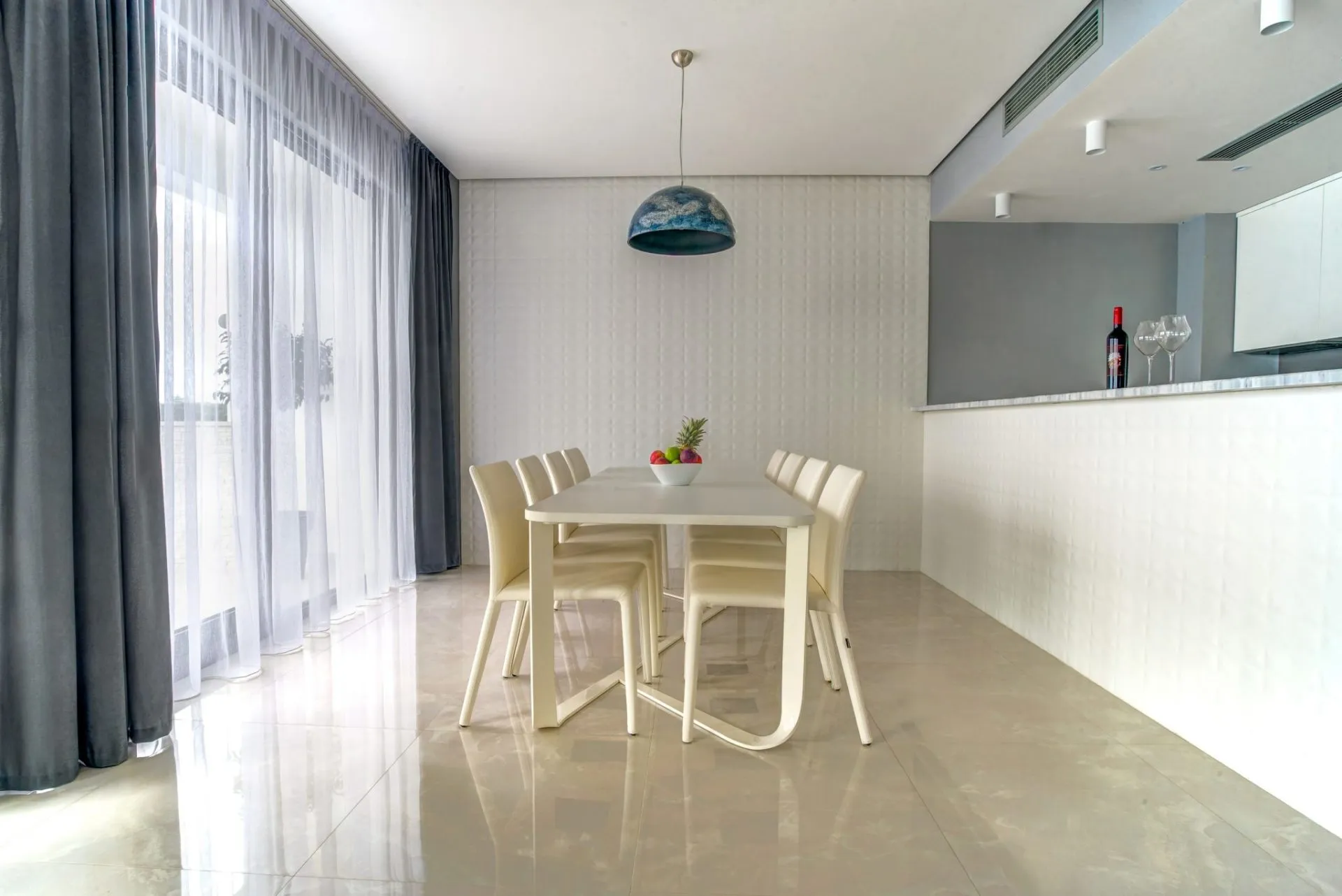 Bright dining area featuring a white table, six cream chairs, and a blue pendant light near sheer curtains.