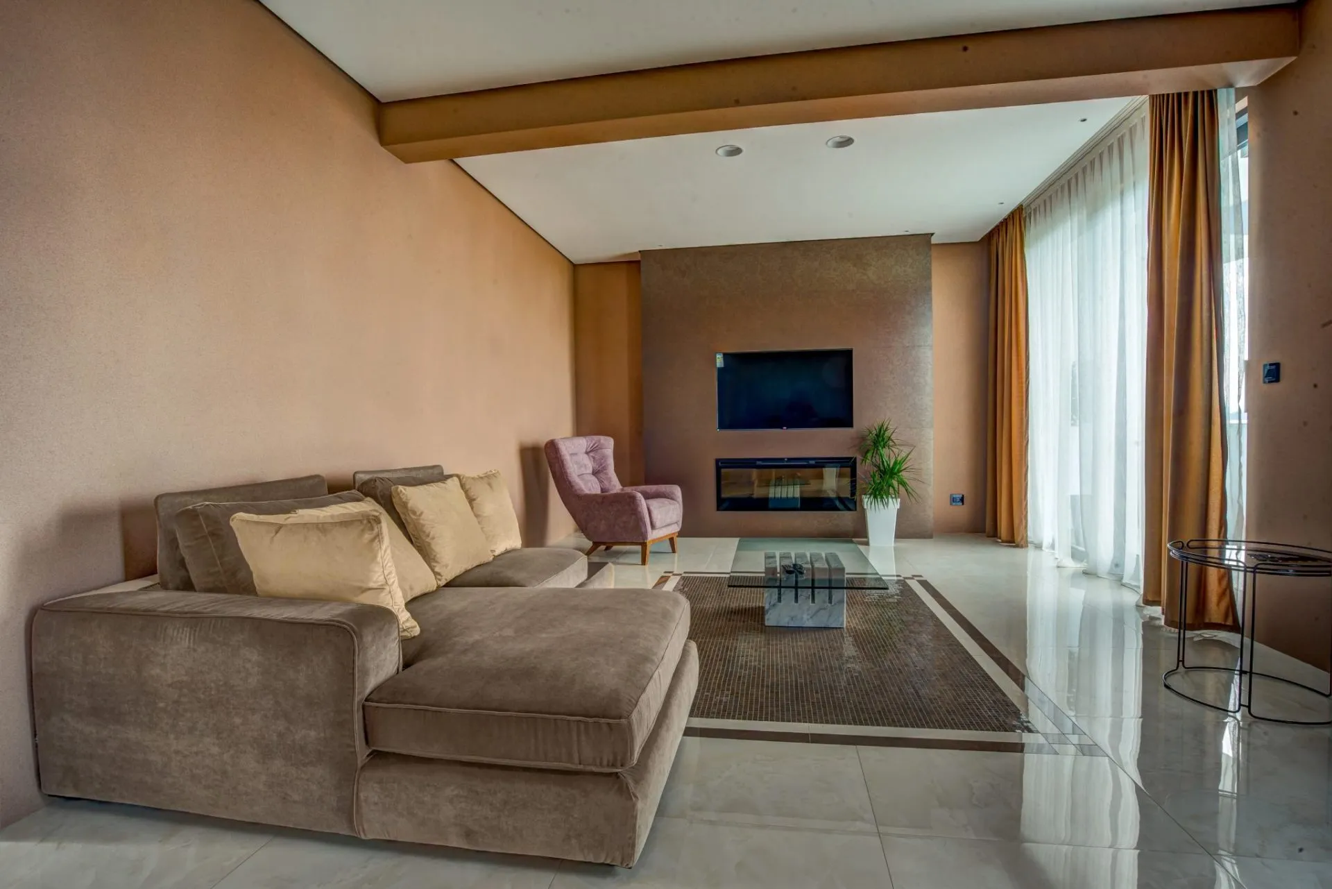 Living room with a brown sectional sofa, wall-mounted TV, fireplace, and pink armchair on glossy tiles.