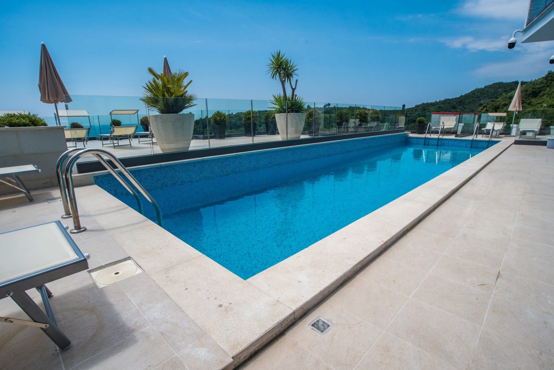 Open-air pool on the villa terrace in Budva, Montenegro, with sun loungers, glass railing, and mountain views.