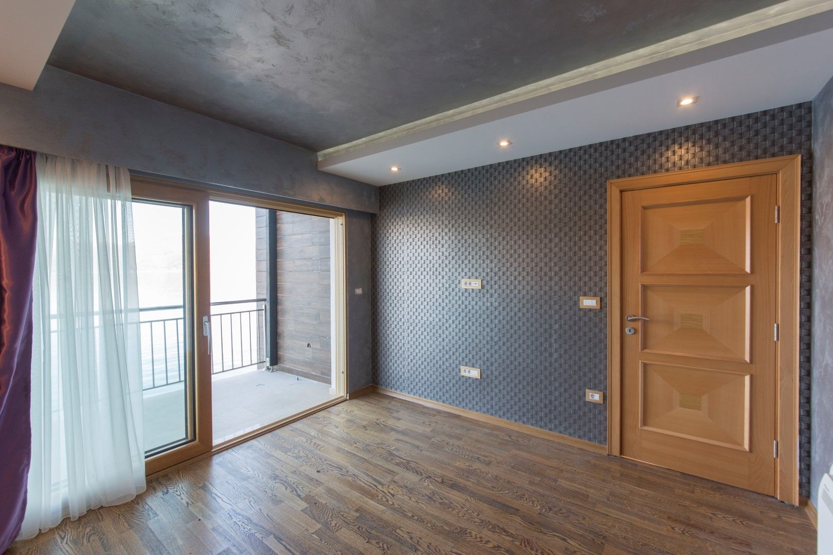 Modern living room with access to the balcony in a villa in Krašići, Montenegro: parquet flooring, textured wallpaper, and a wooden door.