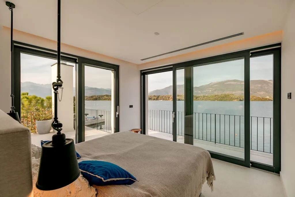 Modern bedroom with a large bed and black pendant lamps. Panoramic windows offer a scenic view of the lake and mountains.