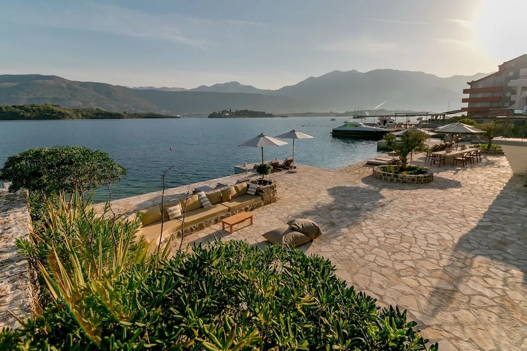 Stone terrace with sofas and umbrellas by the water, mountains and boats in background.