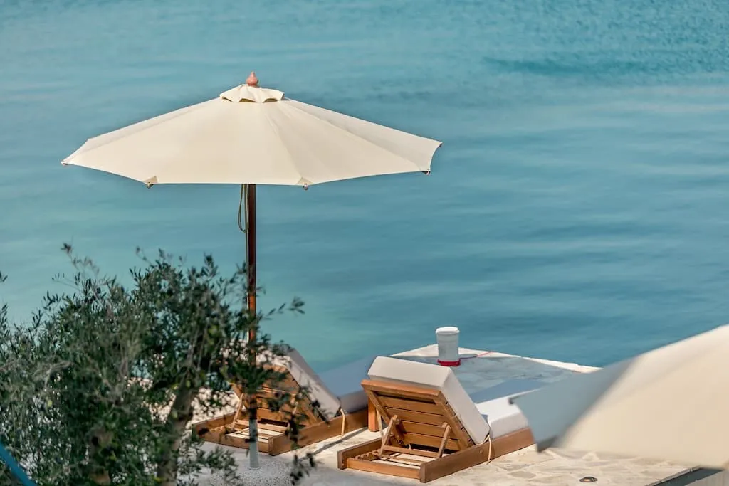Wooden sun loungers and a white umbrella situated by the edge of a pool overlooking blue water.