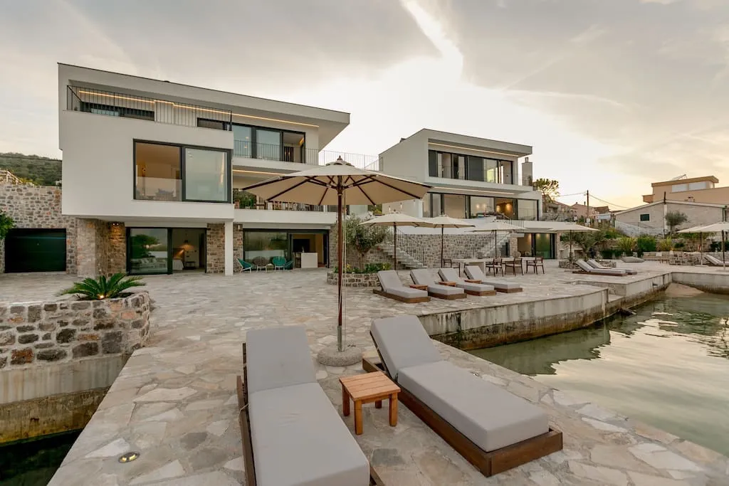 Modern house exterior with stone terrace, sunbeds, umbrellas, and waterfront view at dusk.