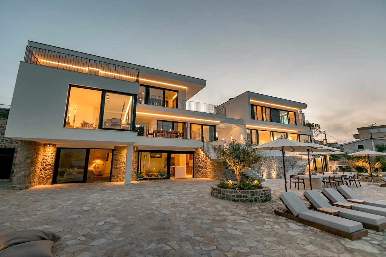 Modern villa in Luštica, Montenegro: panoramic windows, a terrace with sun loungers, stone finishes, and evening lighting.