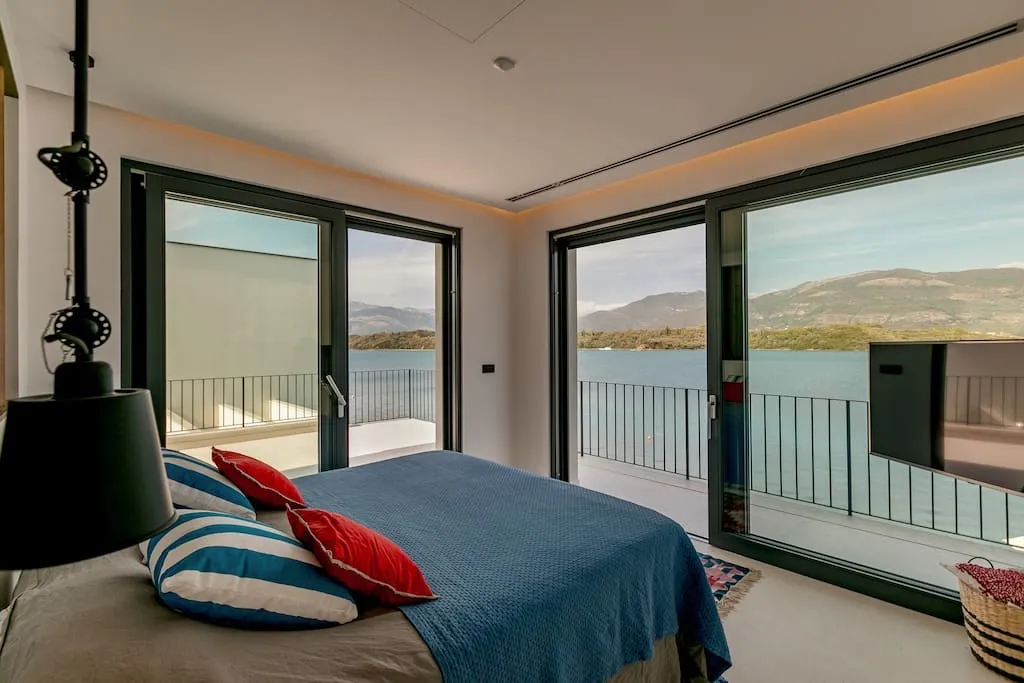 Modern bedroom with a large bed, blue coverlet, and red pillows. Panoramic windows open to a balcony overlooking the sea and mountains.