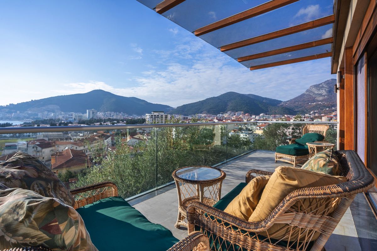 Villa terrace in Budva, Montenegro: wicker furniture, glass railing, and panoramic views of the city and mountains.