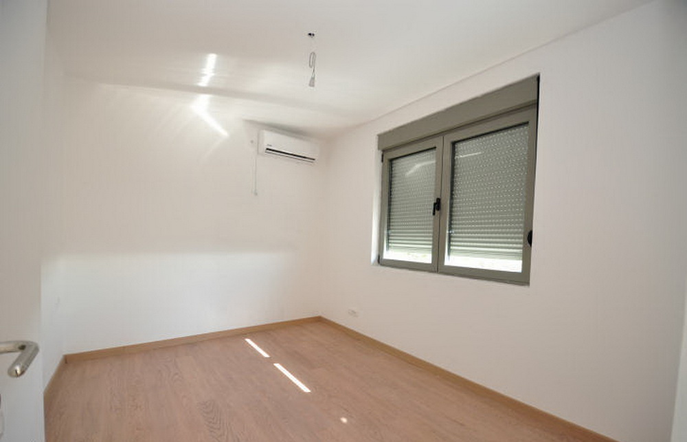 Bright bedroom in a new building in Tivat, Montenegro: laminate flooring, white walls, air conditioner, and a window with roller shutters.