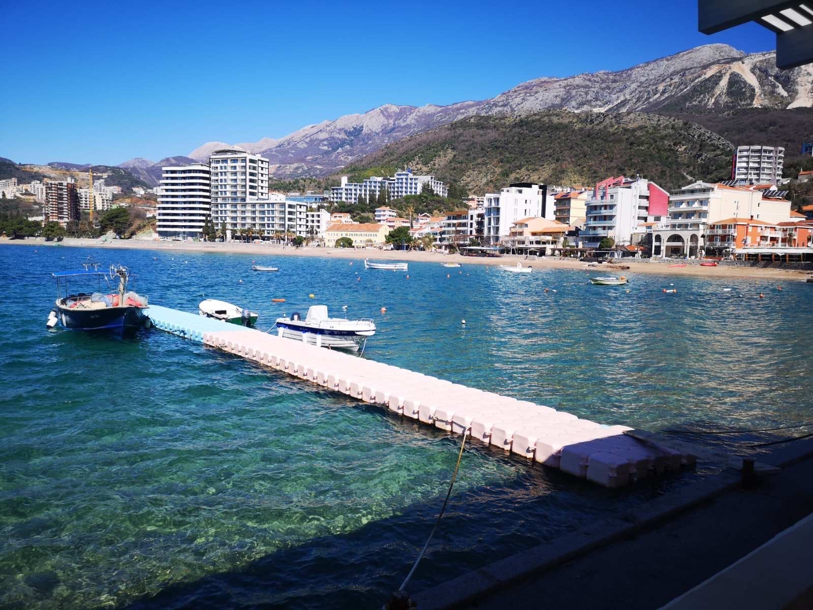 Sea and beach view in Rafailovici, Montenegro: a pier with boats against a backdrop of mountains and the resort infrastructure of the town.