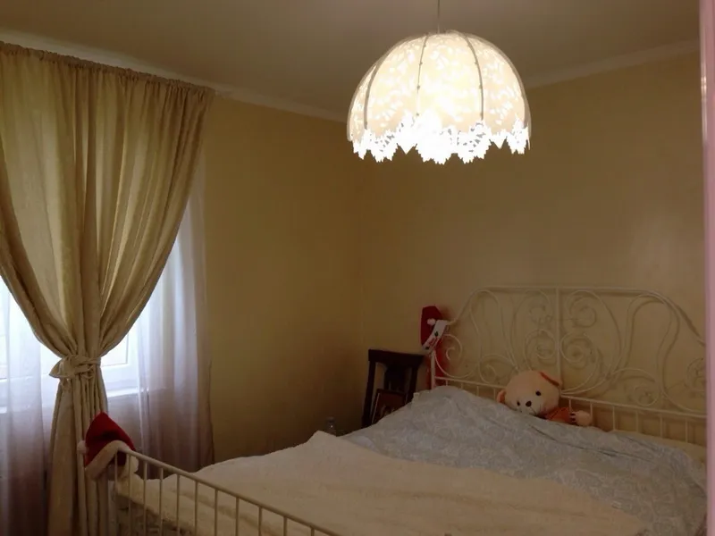 White metal bed frame with teddy bear, lace pendant light, and beige curtains covering a window.