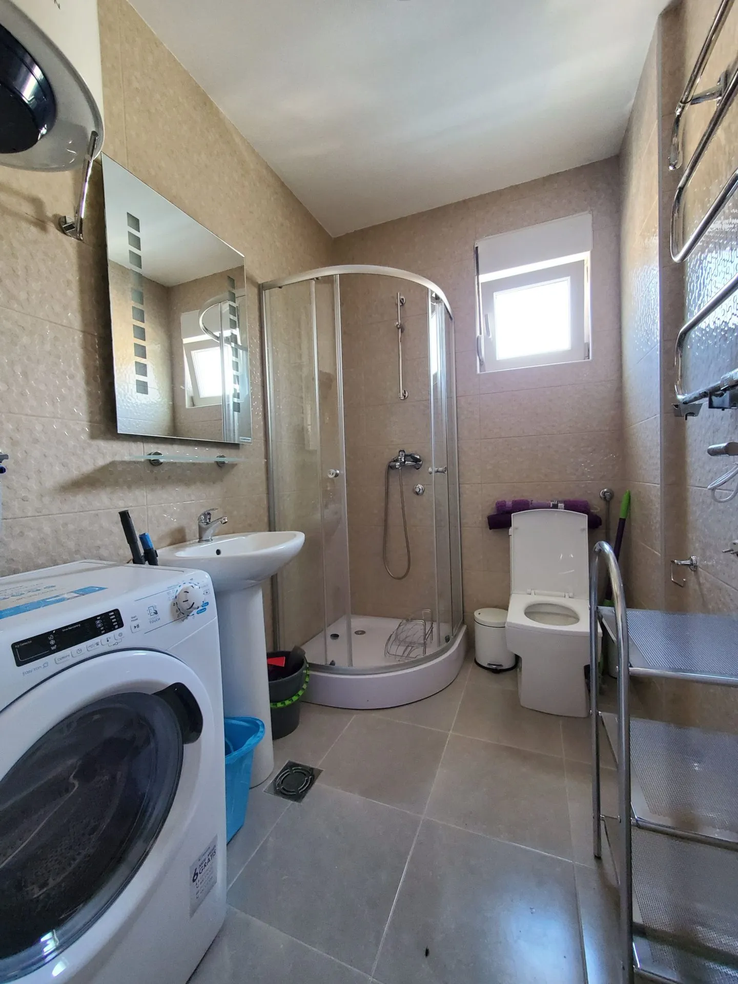 Bathroom featuring a corner shower, toilet, pedestal sink, and washing machine on beige tiles.