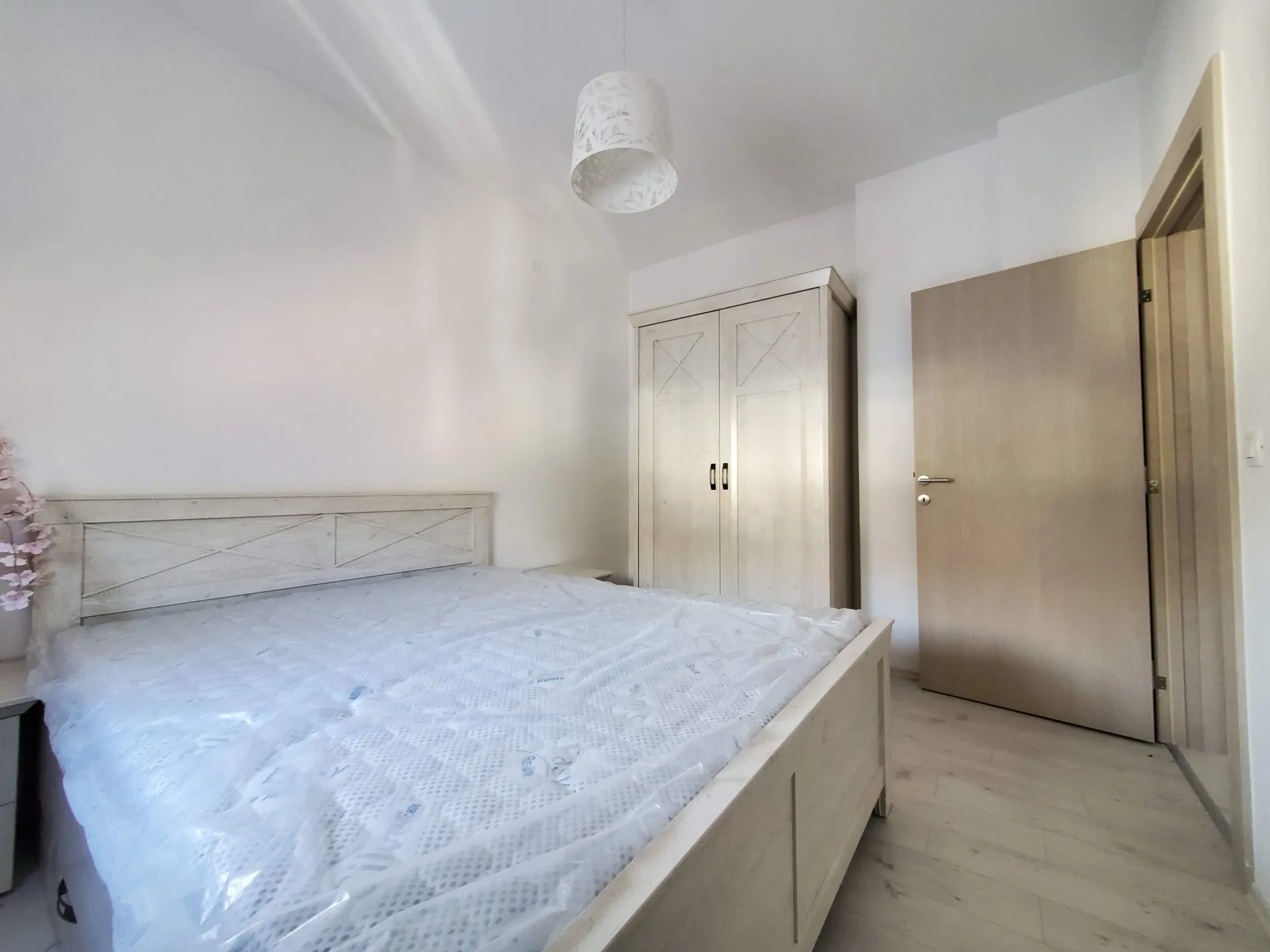 Bright bedroom featuring a wooden bed with plastic-wrapped mattress and a light wardrobe.