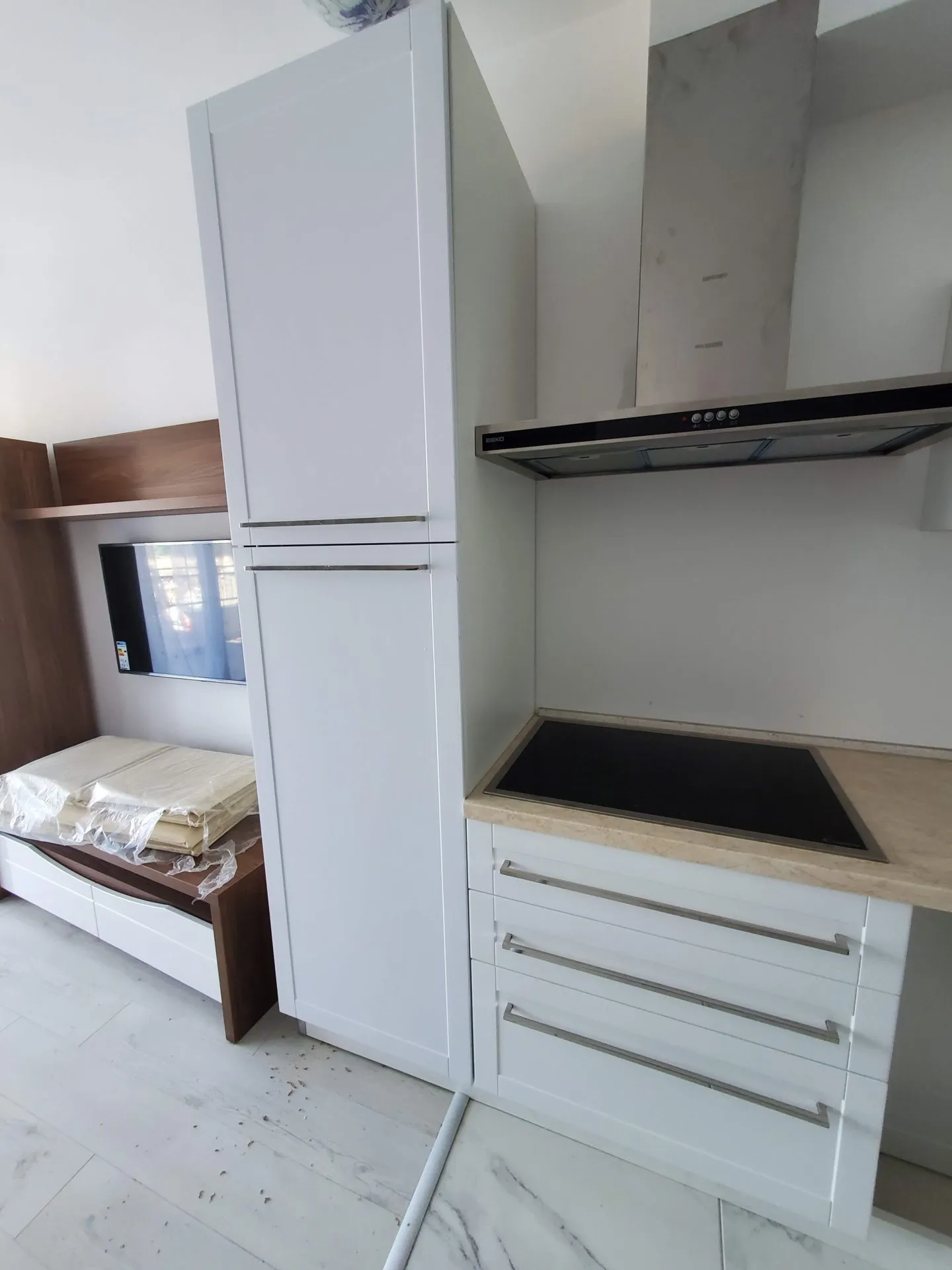 White kitchen cabinets, induction cooktop, stainless steel hood, and TV unit visible.