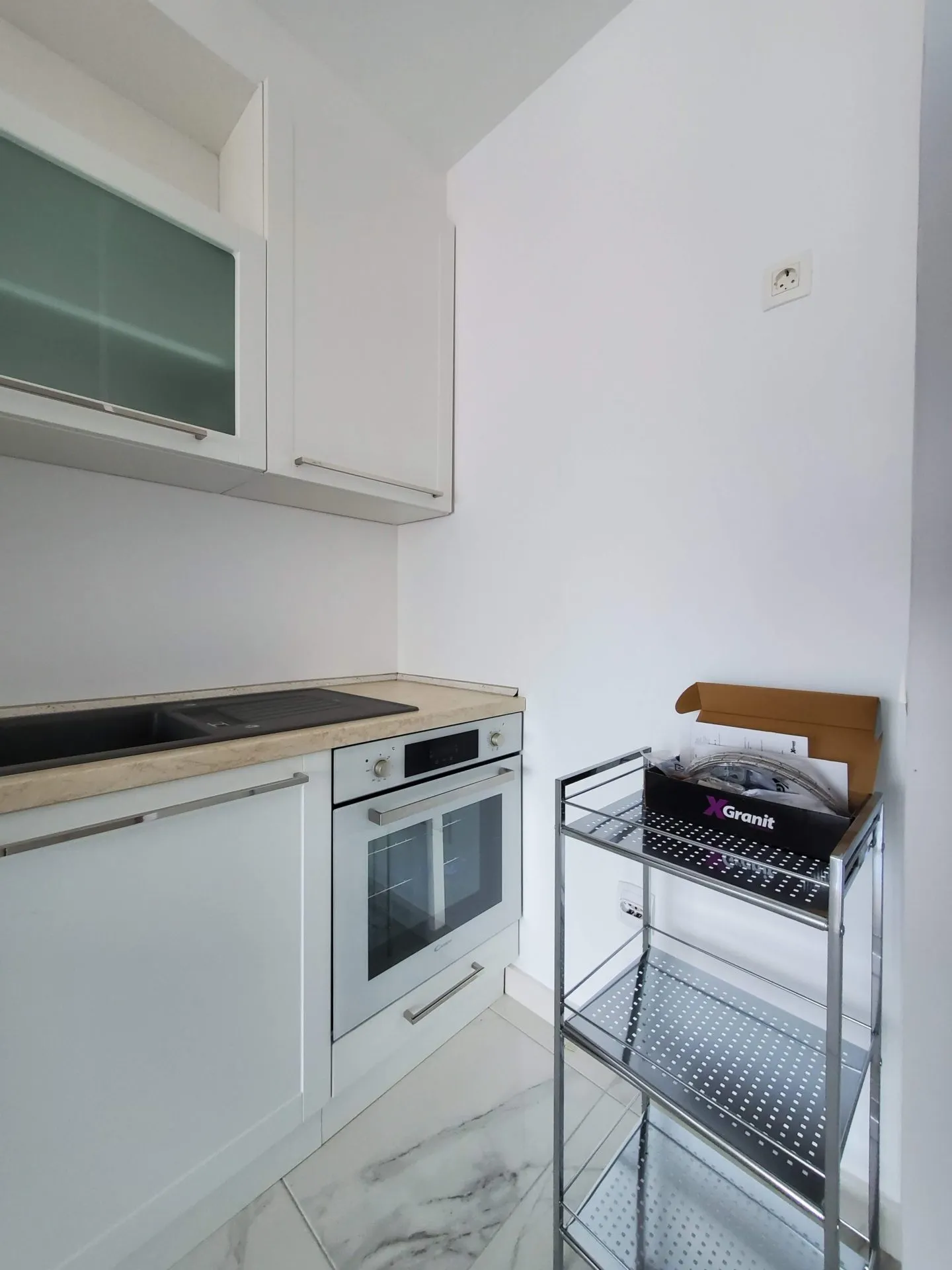 White kitchen cabinets, built-in oven, black sink on a light countertop, and a metal shelving unit.