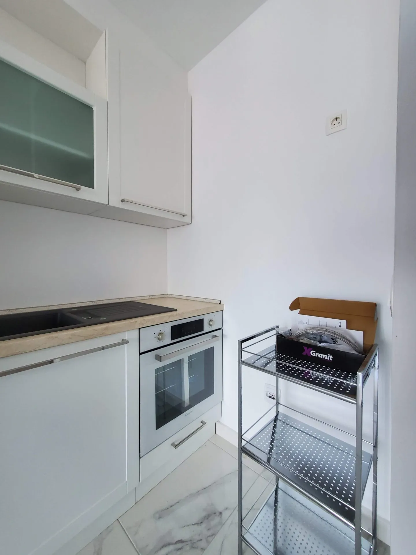 White kitchen with built-in oven, black sink, and metal shelving unit on marble-look tiles.