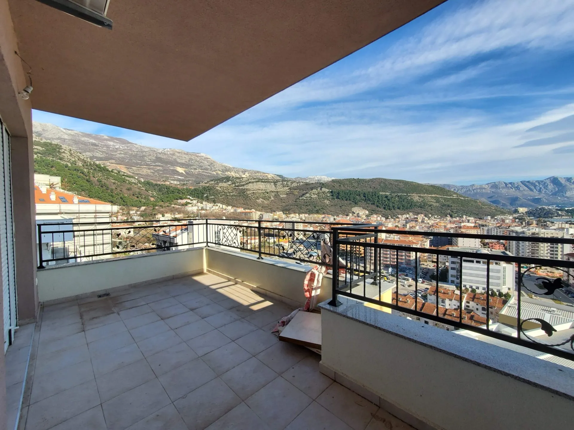 Tiled balcony with ornate iron railing overlooking a cityscape and mountains under a blue sky.