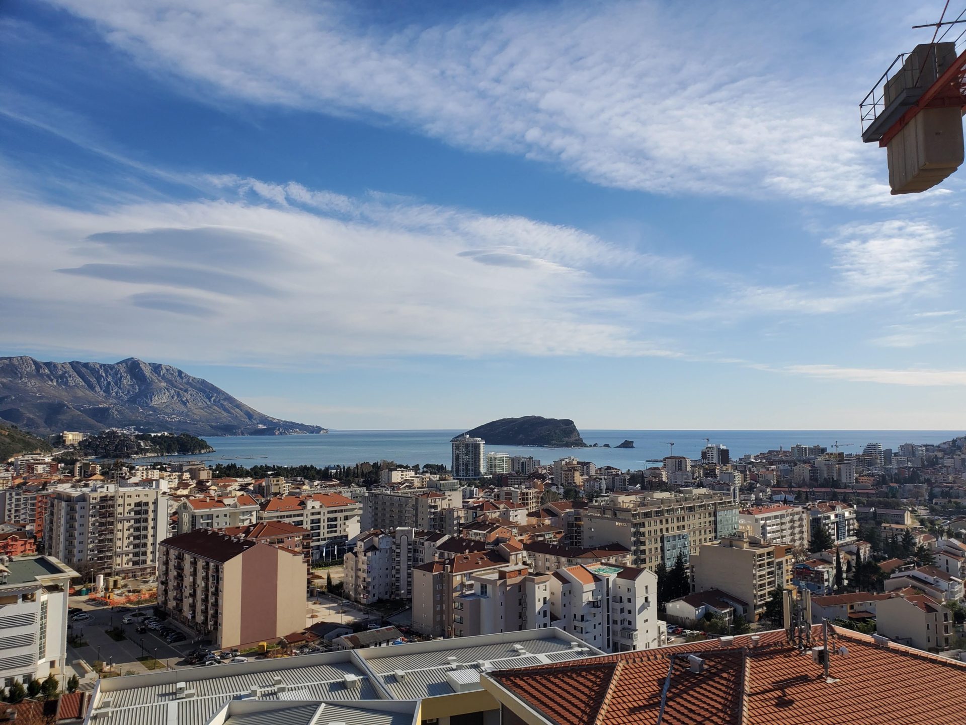 Panoramic view of Budva, the sea, and St. Nicholas Island from the apartment window in Montenegro, with views of the mountains and surrounding development.