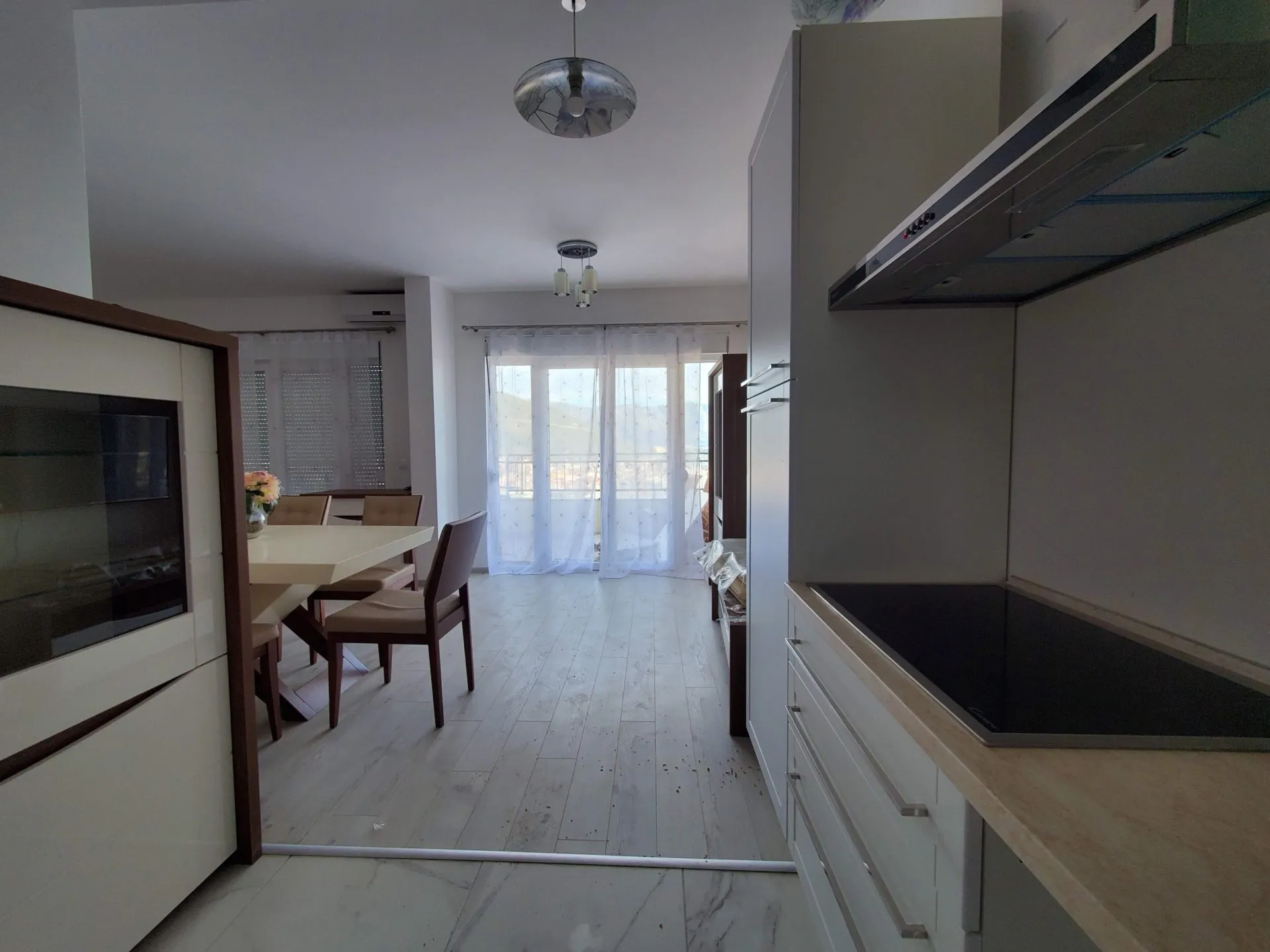 Kitchen area with cooktop and hood, dining table and chairs visible in the background near a window with sheer curtains.