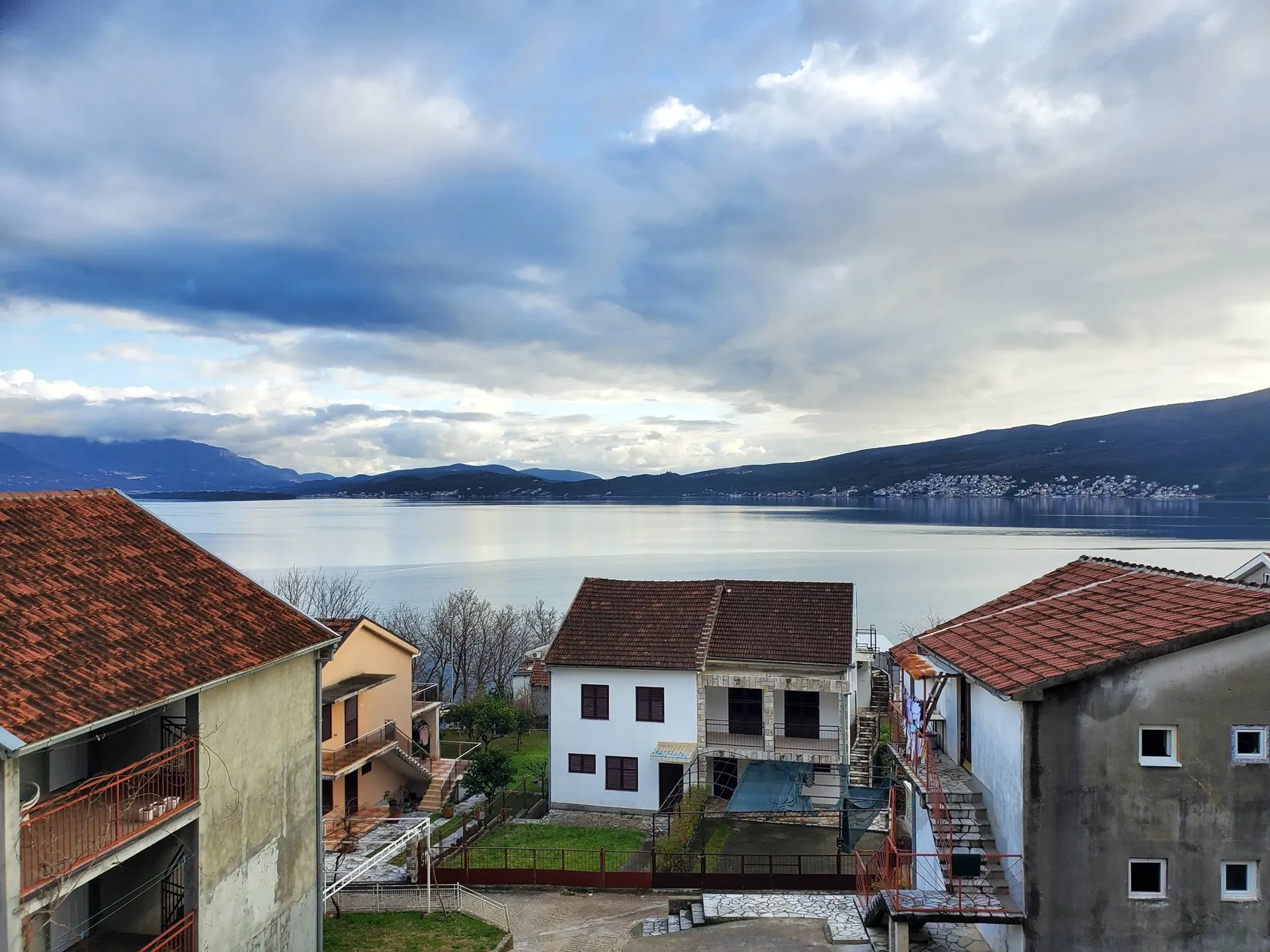 View of houses with red roofs overlooking a calm sea and mountains under a cloudy sky.