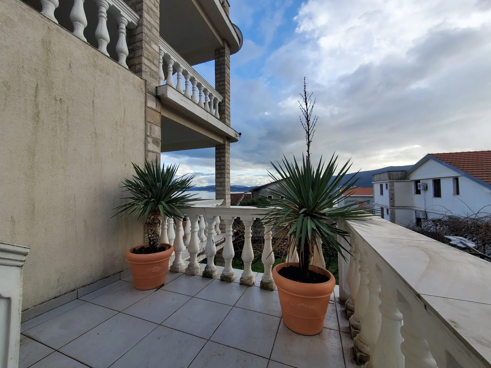 Tiled terrace with white balustrades and two large terracotta pots containing palm-like plants overlooking the sea.