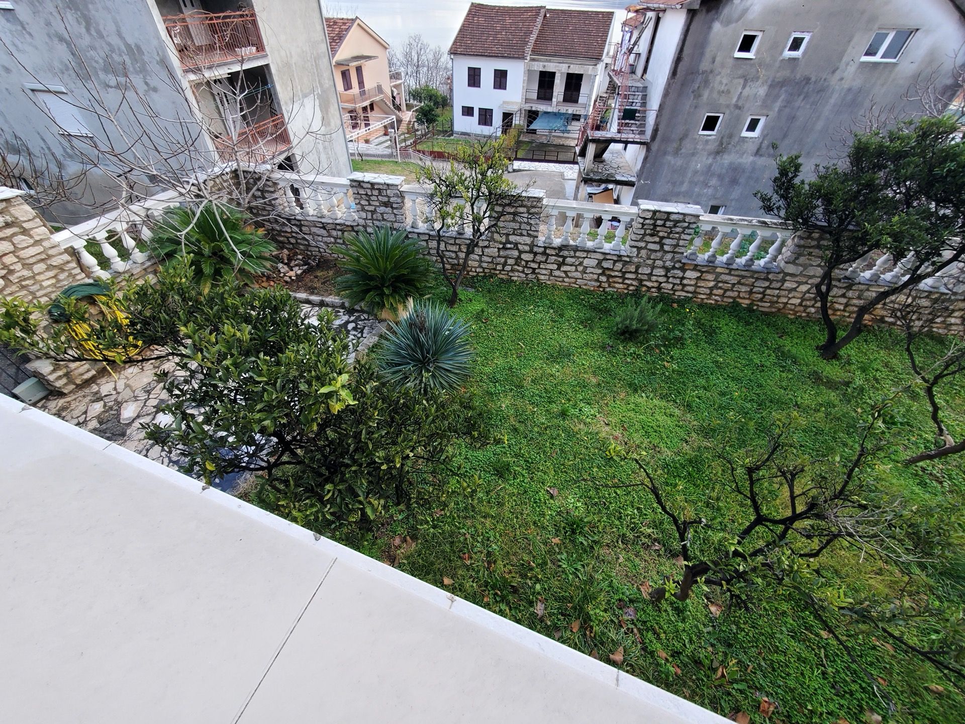 View of the green garden and stone hotel fence in Bijela, Montenegro, from a terrace with white railings and citrus trees.