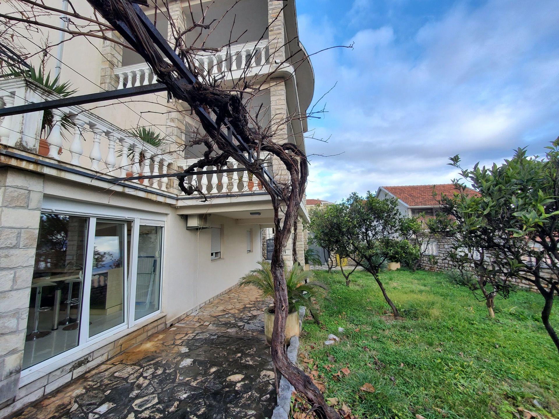 Hotel facade in Bijela, Montenegro: a terrace with stone cladding, a grapevine, and a green garden by the house.