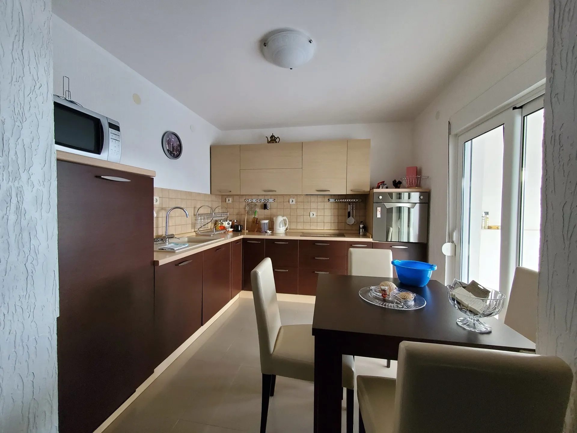 Kitchen with dark lower and light upper cabinets, microwave, sink, oven, and a dining table with chairs near a glass door.