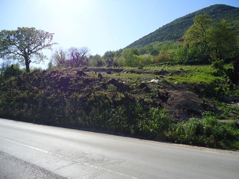 Building plot in Bijela, Montenegro: view of a sloped roadside lot with trees and mountains.