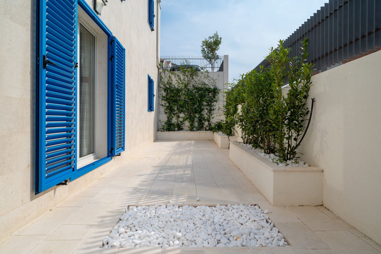 Cozy house terrace in Blizikuće, Montenegro: light tiles, blue shutters, landscaping, and white pebbles.
