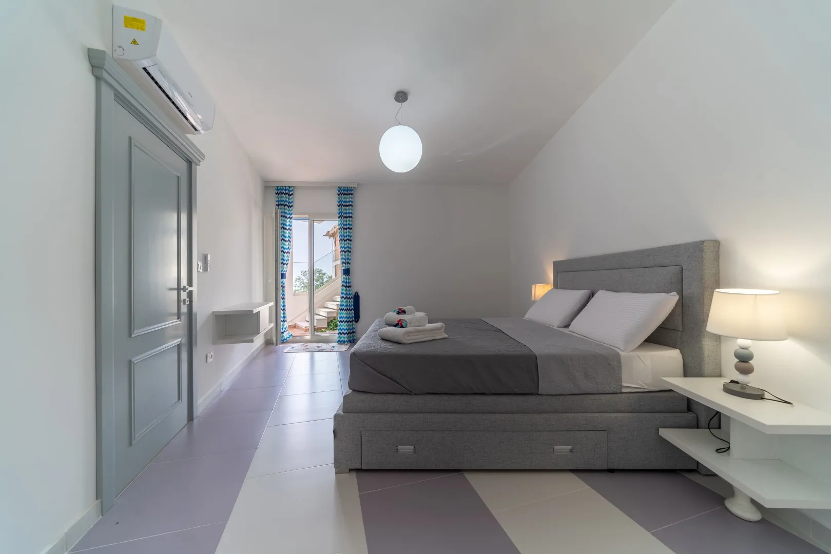 Modern bedroom featuring a grey bed, white nightstands, and a balcony access framed by blue curtains.