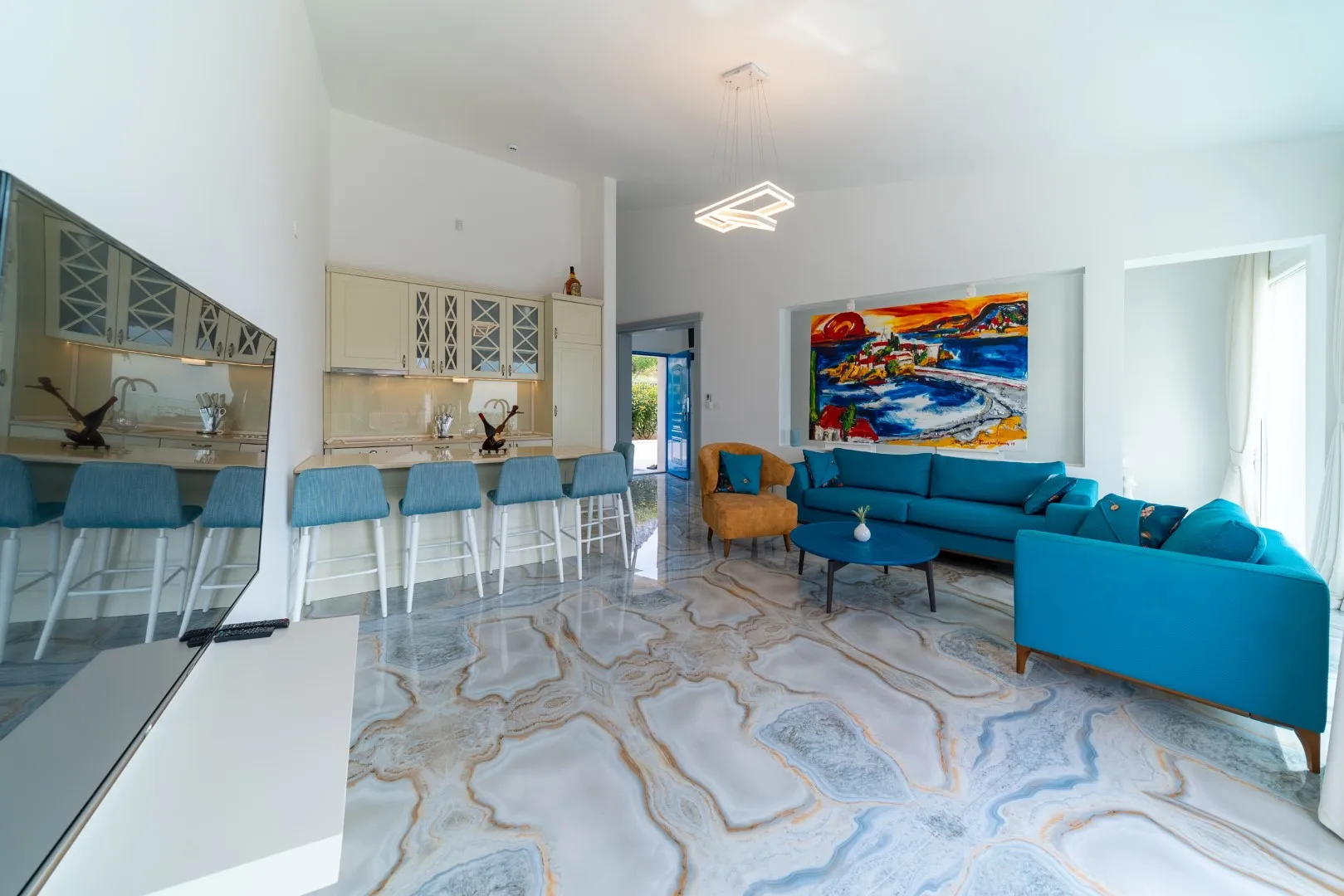 Living room featuring blue sofas, a kitchen bar area with stools, and a colorful painting on patterned floors.
