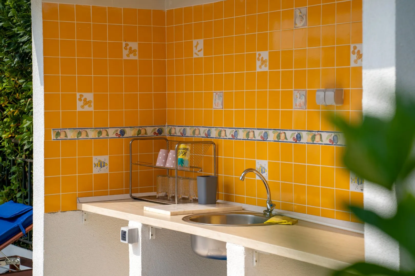 Open kitchen with orange tiles, sink, faucet, and dish rack on a light countertop next to greenery.