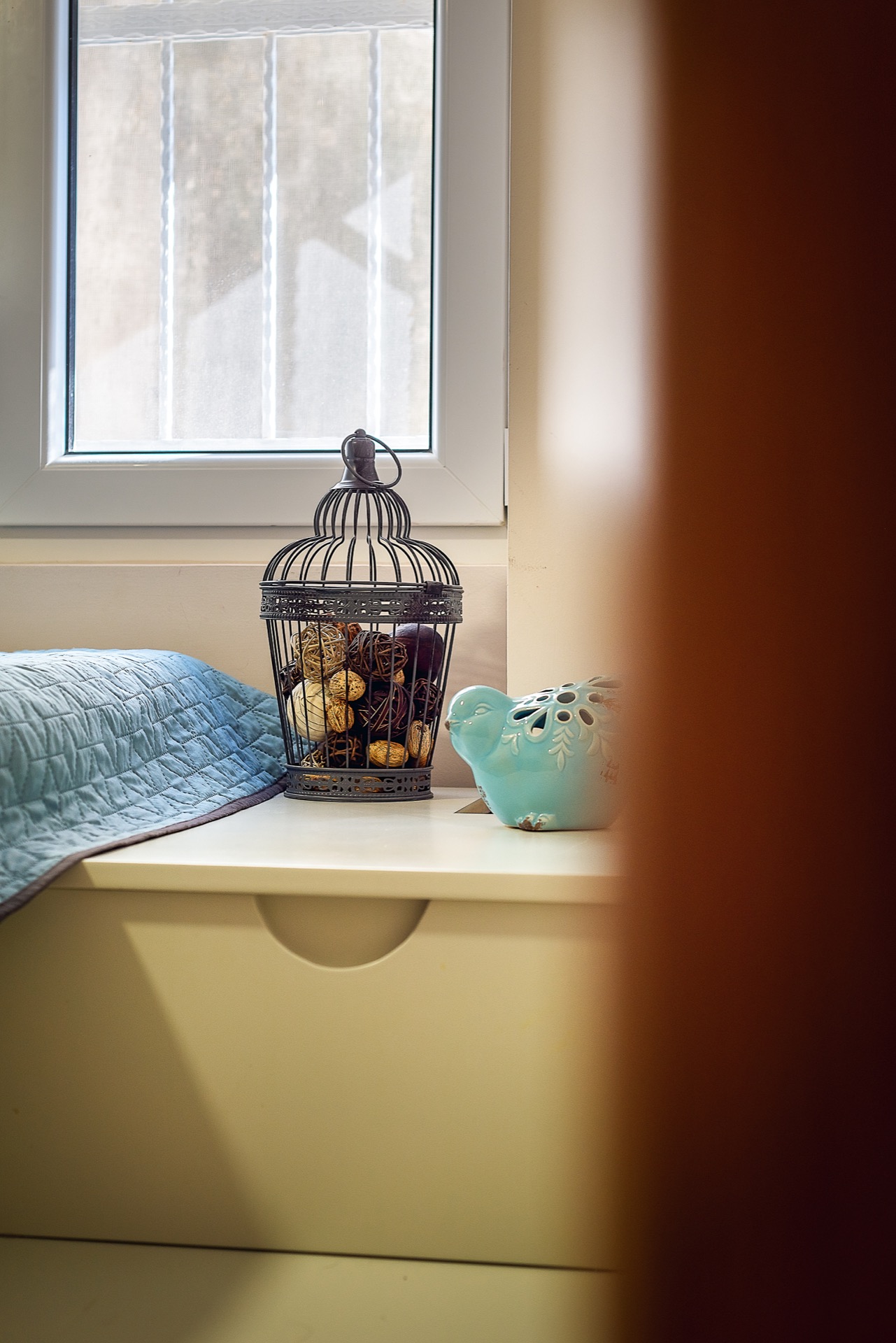 Bedroom decor in an apartment in Budva, Montenegro: a white bedside table, a decorative birdcage, and a bird figurine by the window.