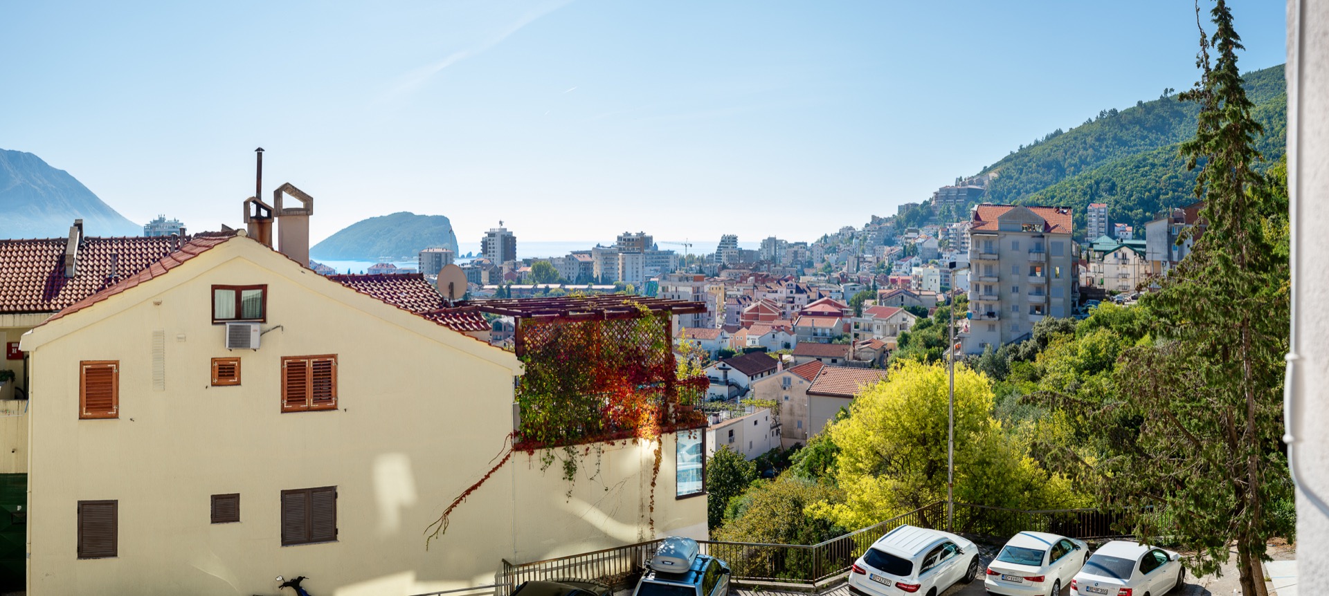 Panoramic view of Budva, Montenegro, with a terrace, green trees, and the sea coast on the horizon.