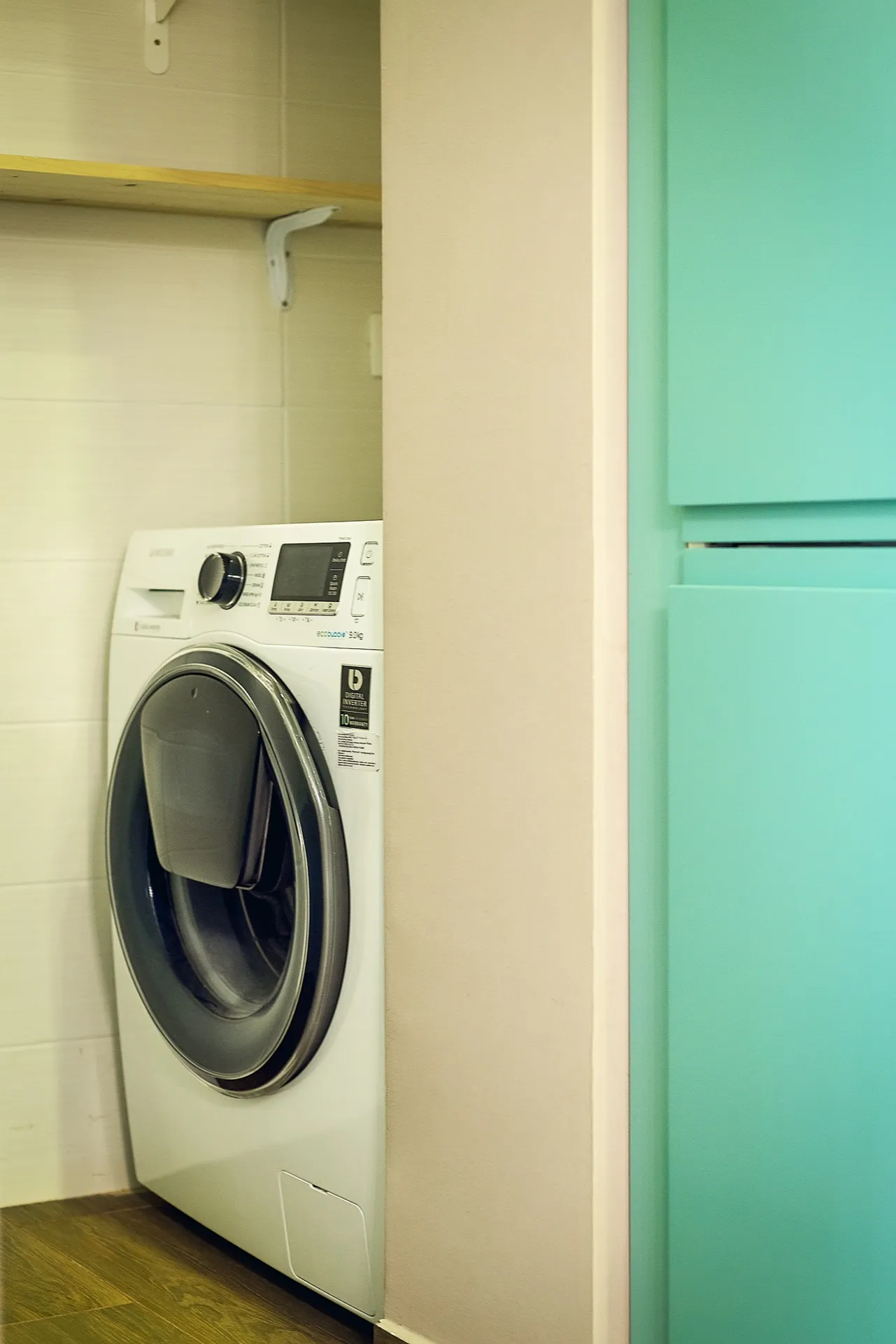 White washing machine in a tiled alcove with a wooden shelf and turquoise cabinet on the right.